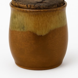 Pode incluir: Um pote de cerâmica com tampa de madeira. O pote é de cor marrom quente com uma faixa verde-oliva perto do topo. A tampa é feita de madeira escura e rachada. O pote tem formato arredondado e está sobre uma superfície branca.