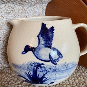 May include: Cream-colored ceramic pitcher with a blue duck design. The duck is depicted in flight above blue water and reeds. The pitcher has a handle and a spout for pouring.
