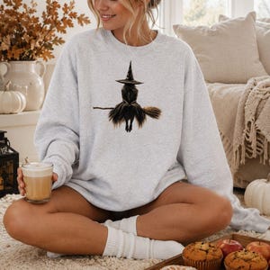 Op de afbeelding: Een lichtgrijs sweatshirt met ronde hals en een centrale grafische illustratie van een heks met een puntmuts die op een bezemsteel zit. De heks is afgebeeld met lang donker haar en een zwierige rok in bruine en zwarte tinten.