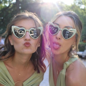 Peut inclure: Deux paires de lunettes de soleil en forme de cœur vert sauge clair avec un texte blanc personnalisé sur les verres. Le verre gauche affiche les initiales M & Q, tandis que le verre droit indique la date 09 08 25 en format numérique.