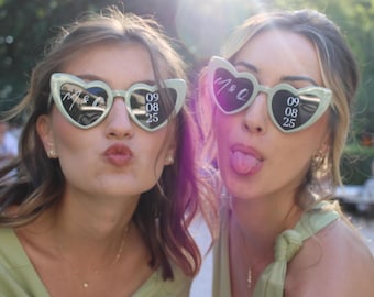 May include: Two pairs of light sage green heart-shaped sunglasses featuring white custom text on the lenses. The left lens displays the initials M & Q, while the right lens shows the date 09 08 25 in a stacked numerical format.