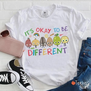 以下が含まれることがあります： 「IT'S OKAY TO BE DIFFERENT」とカラフルな文字で書かれた白いTシャツ。梨、玉ねぎ、ヒマワリの種など、さまざまな食べ物のキャラクターの漫画デザインが特徴です。虹と星もデザインに含まれています。