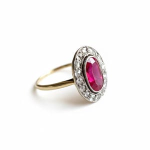 May include: An oval ruby ring with a halo of small diamonds. The ruby is a deep red colour, and the diamonds are clear and sparkling. The ring has a gold band.