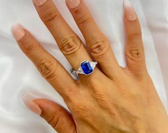 May include: A silver ring featuring a large, rectangular, deep blue gemstone as the centerpiece. Two smaller, clear gemstones flank the main stone. The ring is set on a hand with long, polished nails.