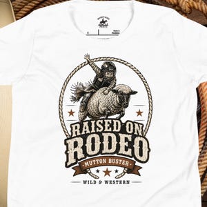 May include: A white short-sleeve t-shirt featuring a graphic of a child in a helmet riding a sheep. The design includes a rope border, stars, and the text RAISED ON RODEO MUTTON BUSTER WILD & WESTERN. The shirt has a Whiskey Ridge Prints label.