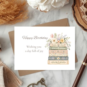 Puede incluir: Una tarjeta de felicitaci&oacute;n rectangular blanca con el texto Happy Birthday y Wishing you a day full of joy. La tarjeta muestra una pila de cinco libros en colores pastel con patrones florales, coronada por un ramo de flores silvestres coloridas.