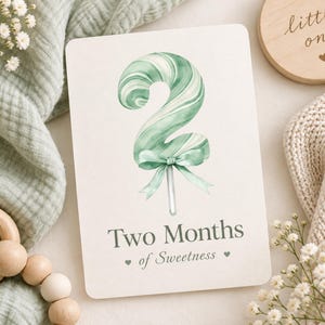 May include: A rectangular milestone card featuring a watercolor illustration of a sage green and white swirled lollipop shaped like the number two, tied with a matching ribbon. The text reads Two Months of Sweetness with small heart icons.