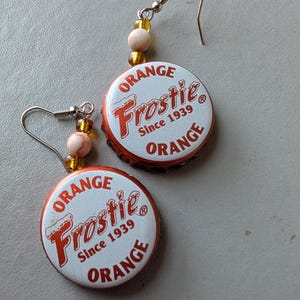 May include: A pair of handmade earrings featuring orange and white bottle caps with the text "ORANGE Frostie Since 1939 ORANGE". The earrings have silver-colored hooks and are accented with small beads.