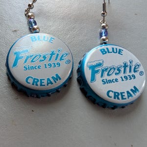 May include: A pair of handmade earrings featuring blue and white bottle caps. The bottle caps are printed with "BLUE Frostie CREAM Since 1939" in blue text. The earrings have silver-colored hooks and are accented with blue and silver beads.