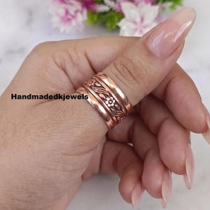 Op de afbeelding: Een brede koperen ring met een centrale band met een in reliëf gemaakt bloemmotief en twee gladde, effen buitenbanden. Het metaal heeft een gepolijste, roodbruine afwerking. De tekst Handmadedkjewels is zichtbaar aan de linkerkant van de ring.