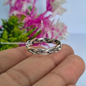 Puede incluir: Un anillo trenzado de color plateado. El anillo tiene un diseño tejido, con múltiples hebras entrelazadas para crear una apariencia texturizada. El anillo se muestra sobre un fondo de flores rosas y verdes.
