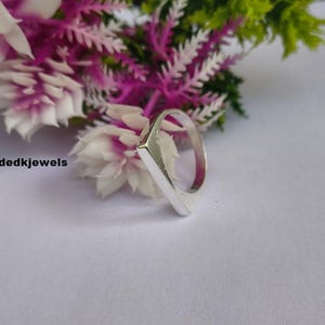 Peut inclure: Une bague en argent avec une face rectangulaire, sur fond de fleurs blanches et violettes. La bague a un design simple et moderne. Le texte "handmadedkjewels" est visible dans l'image.