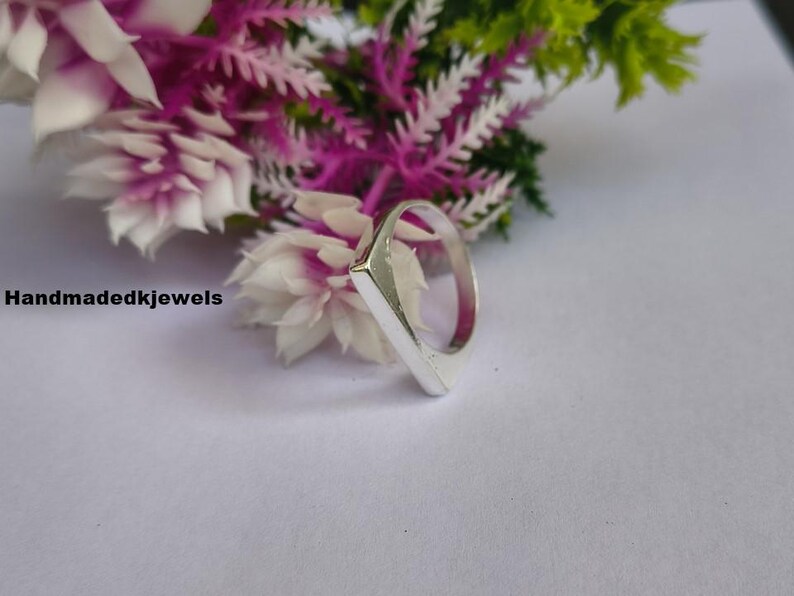 Peut inclure: Une bague en argent avec une face rectangulaire, sur fond de fleurs blanches et violettes. La bague a un design simple et moderne. Le texte "handmadedkjewels" est visible dans l'image.