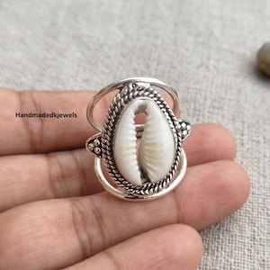 Könnte beinhalten: Ein silberner Ring mit einer zentralen weißen Kaurimuschel, die in eine gedrehte, seilartige Metallfassung eingefasst ist. Der Ring hat ein Doppelband-Design, das sich an den Seiten verbindet und eine offene, geschichtete Optik mit poliertem Finish erzeugt.