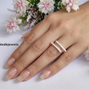 May include: A silver ring featuring two parallel horizontal bars worn on the finger. The design is sleek and metallic with a polished finish. The text Handmadedkjewels is visible on the left side of the image near white and pink flowers.