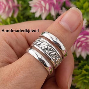 May include: A close-up of a silver ring set on a finger. The ring features a floral design with a polished finish. The text "Handmadedkjewel" is visible in the image. The ring is surrounded by a blurred background of pink and green flowers.