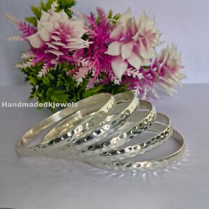 May include: A collection of silver bangles with a hammered finish, stacked together. The bangles are presented against a backdrop of pink and white flowers and green leaves. The text "Handmadekjewels" is visible in the image.