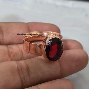 May include: A copper ring featuring a large, oval-cut, deep red garnet gemstone set in a hammered metal band. The band has a wide, textured surface with a metallic sheen. The text Handmadedkjewels is visible on the side of the ring band.