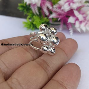 Puede incluir: Un anillo de plata con un diseño único, con múltiples elementos esféricos. El anillo tiene varias bandas y esferas de diferentes tamaños. El anillo se muestra en una mano, con un fondo floral borroso.