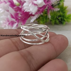 May include: A silver ring with a unique, interwoven design. The ring features multiple bands and crisscrossing lines, creating an abstract and modern look. The ring is shown against a blurred background of pink and green flowers.