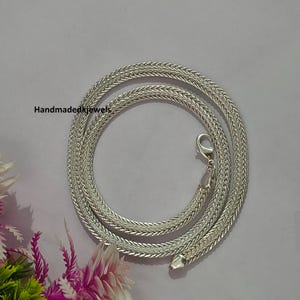 May include: A silver-coloured chain necklace featuring a tightly woven, braided pattern with a metallic sheen. The necklace is arranged in a circular coil and includes a lobster claw clasp. The text "Handmadedkjewels" is printed in black on the left.