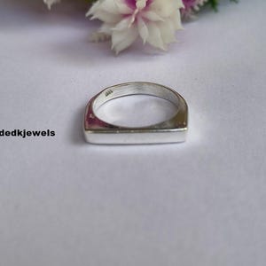 May include: A silver ring with a rectangular face. The ring is set against a white background with a few flowers in the background. The text "Handmadekjewels" is on the left side of the image.