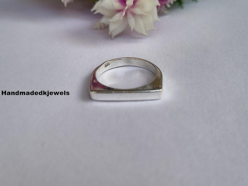 Peut inclure: Une bague en argent avec une face rectangulaire. La bague est pos&eacute;e sur un fond blanc avec quelques fleurs en arri&egrave;re-plan. Le texte "Handmadekjewels" est sur le c&ocirc;t&eacute; gauche de l'image.