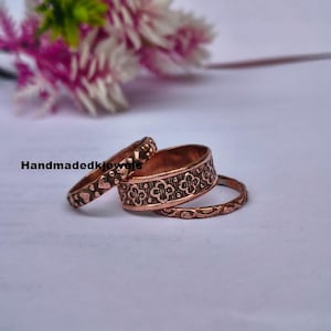 May include: A set of three copper-toned rings featuring distinct textures and patterns. One ring displays a repeating quatrefoil motif, another has a textured surface with small raised dots, and the third is a thin band with a subtle embossed design.