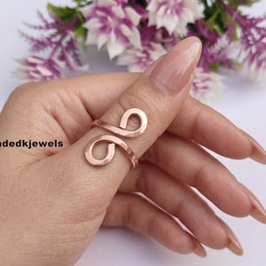 May include: A rose gold-toned metal ring featuring an open-ended design with two symmetrical, rounded loops that curve inward. The ring is worn on a thumb and displays the text Handmadedkjewels on the left side of the image.