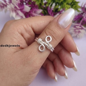Könnte beinhalten: Ein silberfarbener Drahtring mit einem zentralen Knotendesign, zwei geschlungenen Enden und gewickelten Drahtdetails am Ringband. Der Text 'Handmadedkjewels' ist auf der linken Seite des Bildes in der Nähe des Rings zu sehen.