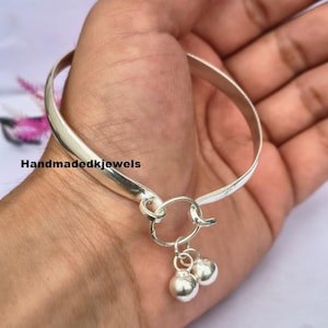 May include: A polished silver-tone bangle bracelet featuring a smooth, open-ended band that hooks together with a circular clasp. Two small, round, metallic silver-tone charms hang from the central ring closure. The text Handmadekjewels is visible.