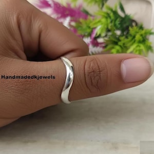 May include: A silver ring with a curved design. The ring is worn on a finger, with the text "Handmadekjewels" visible. The background features blurred greenery and flowers.