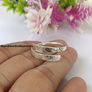 May include: A silver wrap ring with a hammered texture and floral details. The ring is shown on a hand, with the text "Handmadekjewels" visible. The ring's design features an overlapping spiral.