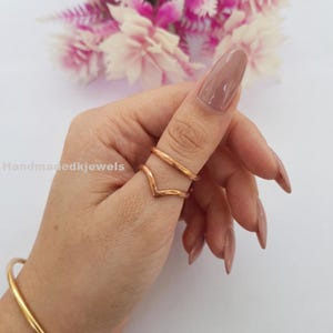May include: Two copper-colored rings are displayed on a finger. The top ring is a simple band, while the lower ring forms a V-shape. The background includes a blurred arrangement of pink and white flowers and the text "Handmadekjewels".