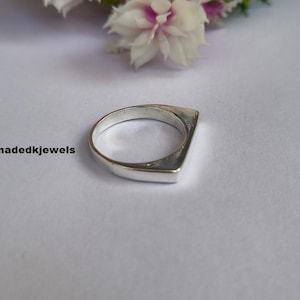 Peut inclure: Une bague en argent au design g&eacute;om&eacute;trique. La bague a un dessus plat et rectangulaire et un anneau arrondi. Le texte "Handmadekjewels" est visible dans l'image. La bague est pr&eacute;sent&eacute;e sur une surface blanche avec des fleurs en arri&egrave;re-plan.