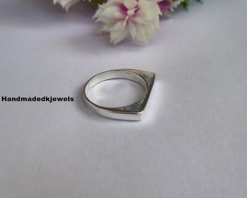 Peut inclure: Une bague en argent au design g&eacute;om&eacute;trique. La bague a un dessus plat et rectangulaire et un anneau arrondi. Le texte "Handmadekjewels" est visible dans l'image. La bague est pr&eacute;sent&eacute;e sur une surface blanche avec des fleurs en arri&egrave;re-plan.