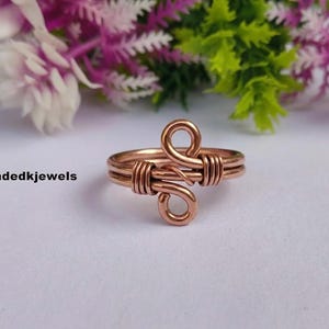 May include: A copper wire ring featuring a central design of two mirrored loops connected by wrapped wire coils. The band is constructed from multiple strands of polished copper wire. The text Handmadedkjewels is visible on the left side.