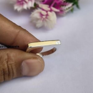 Peut inclure: Une bague en argent avec un dessus rectangulaire de couleur or. La bague est tenue par une main sur un fond doux et clair. Le texte "Handmadekjewels" est visible sur le c&ocirc;t&eacute; gauche de l'image.