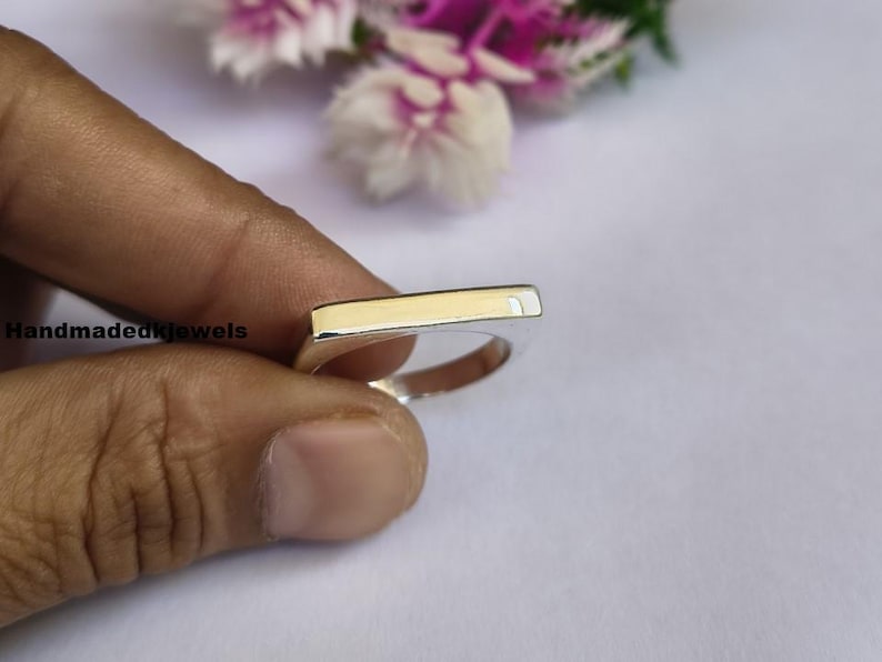 Peut inclure: Une bague en argent avec un dessus rectangulaire de couleur or. La bague est tenue par une main sur un fond doux et clair. Le texte "Handmadekjewels" est visible sur le c&ocirc;t&eacute; gauche de l'image.