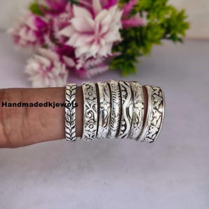 Puede incluir: Una colección de anillos de plata con intrincados diseños florales y de hojas. Los anillos se muestran en un dedo, con un fondo floral borroso. Los anillos están grabados con varios diseños, mostrando la artesanía.