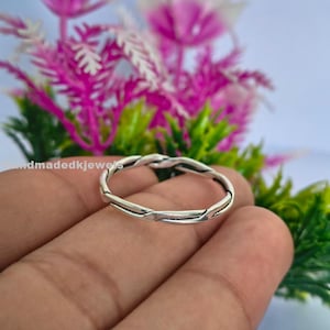 May include: A silver-colored ring with a twisted rope design. The ring is held in a hand, with a blurred background of pink and green foliage. The ring is a simple band.