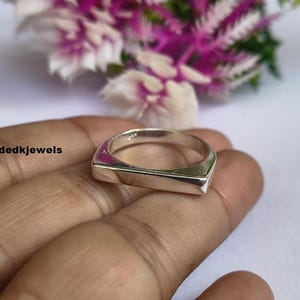 Peut inclure: Une bague carr&eacute;e argent&eacute;e avec une surface et des c&ocirc;t&eacute;s plats. La bague est tenue dans une main, avec un arri&egrave;re-plan flou de fleurs roses et blanches. Le texte "Handmadedkjewels" est visible sur le c&ocirc;t&eacute; gauche.