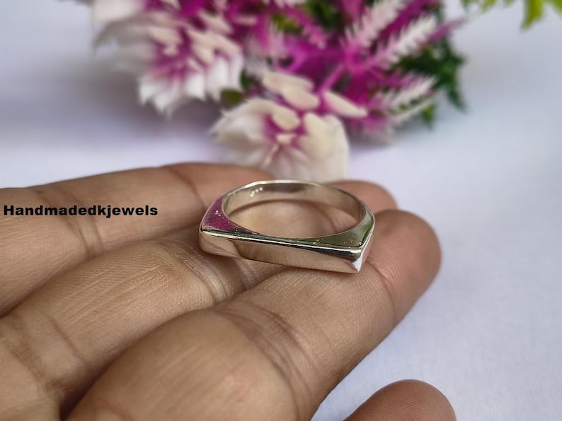 Peut inclure: Une bague carr&eacute;e argent&eacute;e avec une surface et des c&ocirc;t&eacute;s plats. La bague est tenue dans une main, avec un arri&egrave;re-plan flou de fleurs roses et blanches. Le texte "Handmadedkjewels" est visible sur le c&ocirc;t&eacute; gauche.