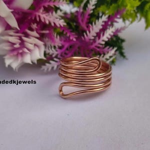 May include: A copper-colored wire ring featuring a multi-layered spiral design with rounded ends. The ring is positioned on a plain surface with the text Handmadedkjewels visible to the left. The metal has a smooth, polished finish.