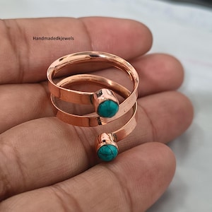 Peut inclure: Une bague en cuivre de type bypass avec deux extrémités ouvertes, chacune surmontée d'une petite pierre ronde de couleur turquoise. L'anneau métallique présente une finition polie et réfléchissante. Le texte "Handmadedkjewels" est visible.