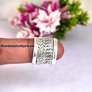 Könnte beinhalten: Ein silberner Ring mit einem strukturierten Design, das eine Reihe miteinander verbundener Bänder aufweist. Der Ring hat ein detailliertes Muster, das gewebten Blättern ähnelt. Der Hintergrund umfasst eine verschwommene Anordnung von rosa und grünen Blumen, mit dem Text "Handmadedkjewels".