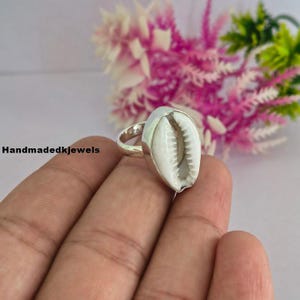 May include: A silver ring featuring a white cowrie shell as the centerpiece. The shell has a smooth, oval shape with a central slit and ribbed edges. The metal band is polished and holds the shell securely in place. The text Handmadekjewels is visible.