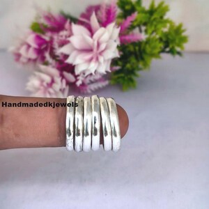 May include: A collection of silver rings, stacked on a finger. The rings have a hammered texture and a shiny finish. The background features a blurred arrangement of pink, white, and green flowers. The text "handmadedkjewels" is visible.