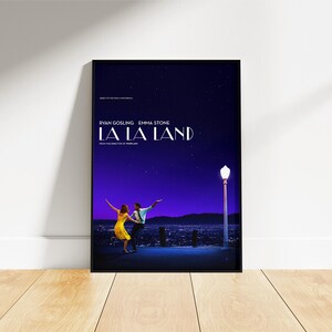 May include: A framed movie poster for La La Land featuring a dark blue starry night sky, a city skyline, and a street lamp. A person in a yellow dress and a person in a white shirt and dark trousers are dancing. Text includes the title and cast names.