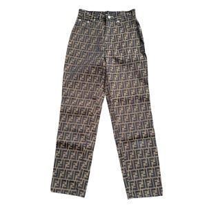 May include: High-waisted brown and black patterned trousers. The trousers feature a repeating logo design. They have a button and zip closure and belt loops. The trousers are made of a woven fabric.